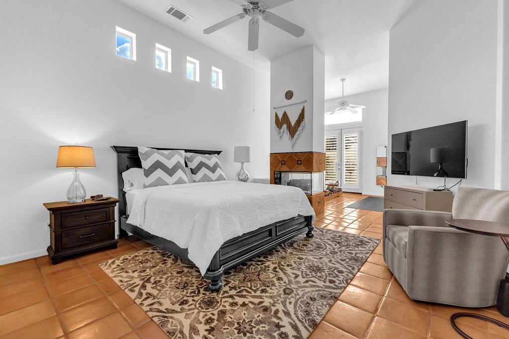 A bedroom with a large bed, nightstands with lamps, a mounted TV, a cozy armchair, and a fireplace. The room has white walls, a tiled floor, and small high windows.