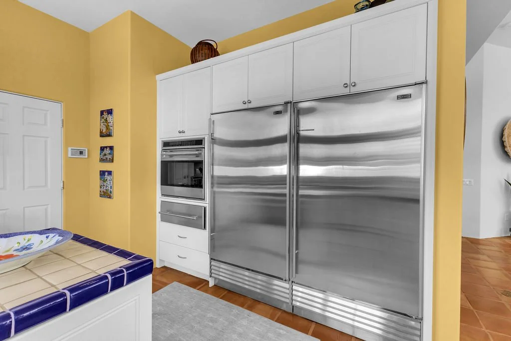 Kitchen with yellow walls, white cabinetry, stainless steel double-door refrigerator, built-in microwave and oven, partially visible tiled table, and small decorative art on the wall.