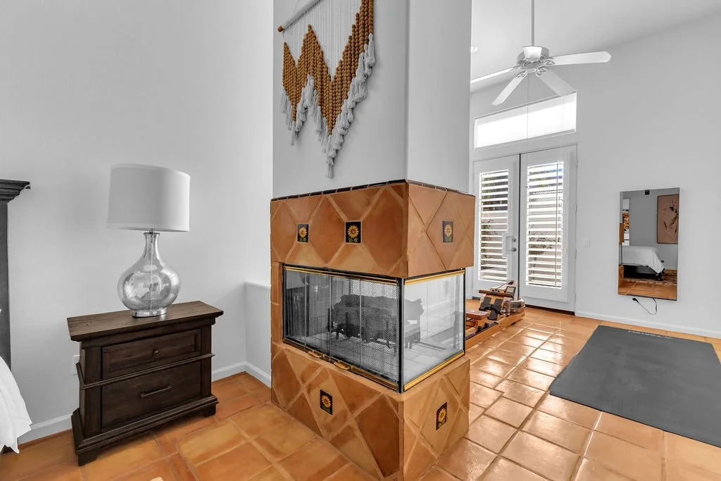 Interior of a living space featuring a brick fireplace with a glass enclosure, a dark wood side table with a glass lamp, a gray rug, a mirror on the wall, and a door leading outside with white shutters.