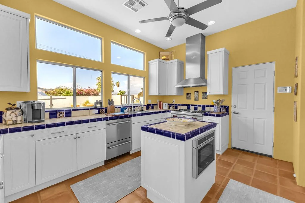 Bright kitchen with yellow walls, white cabinets, blue tiled countertops, and a small island with an oven. Large windows let in natural light, a stainless steel range hood is above the stove, and a ceiling fan is overhead.