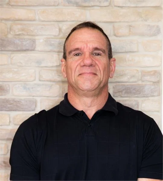 A middle-aged man standing in front of a brick wall, wearing a black polo shirt, with arms crossed and a neutral expression.