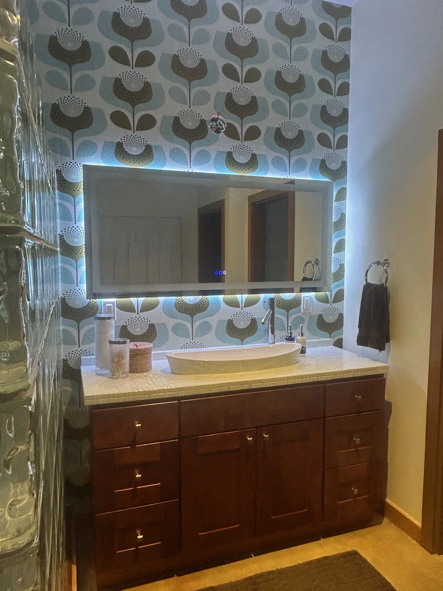 Bathroom vanity with a large mirror, brown wooden cabinet, tiled countertop, a white basin sink, soap dispenser, small basket, and decorative items. The wall above the vanity has patterned wallpaper with stylized flowers and leaves, with LED lighting