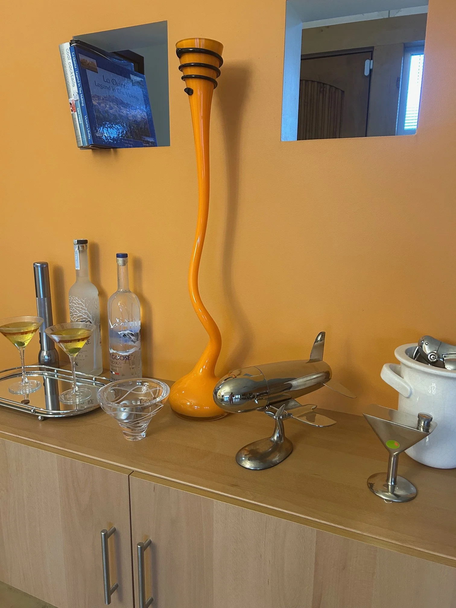 A kitchen counter with various barware and decorative items, including a tall yellow and black glass sculpture, two bottles of liquor, martini glasses, a metal wine opener, a white ceramic bowl, and a decorative metal airplane sculpture.