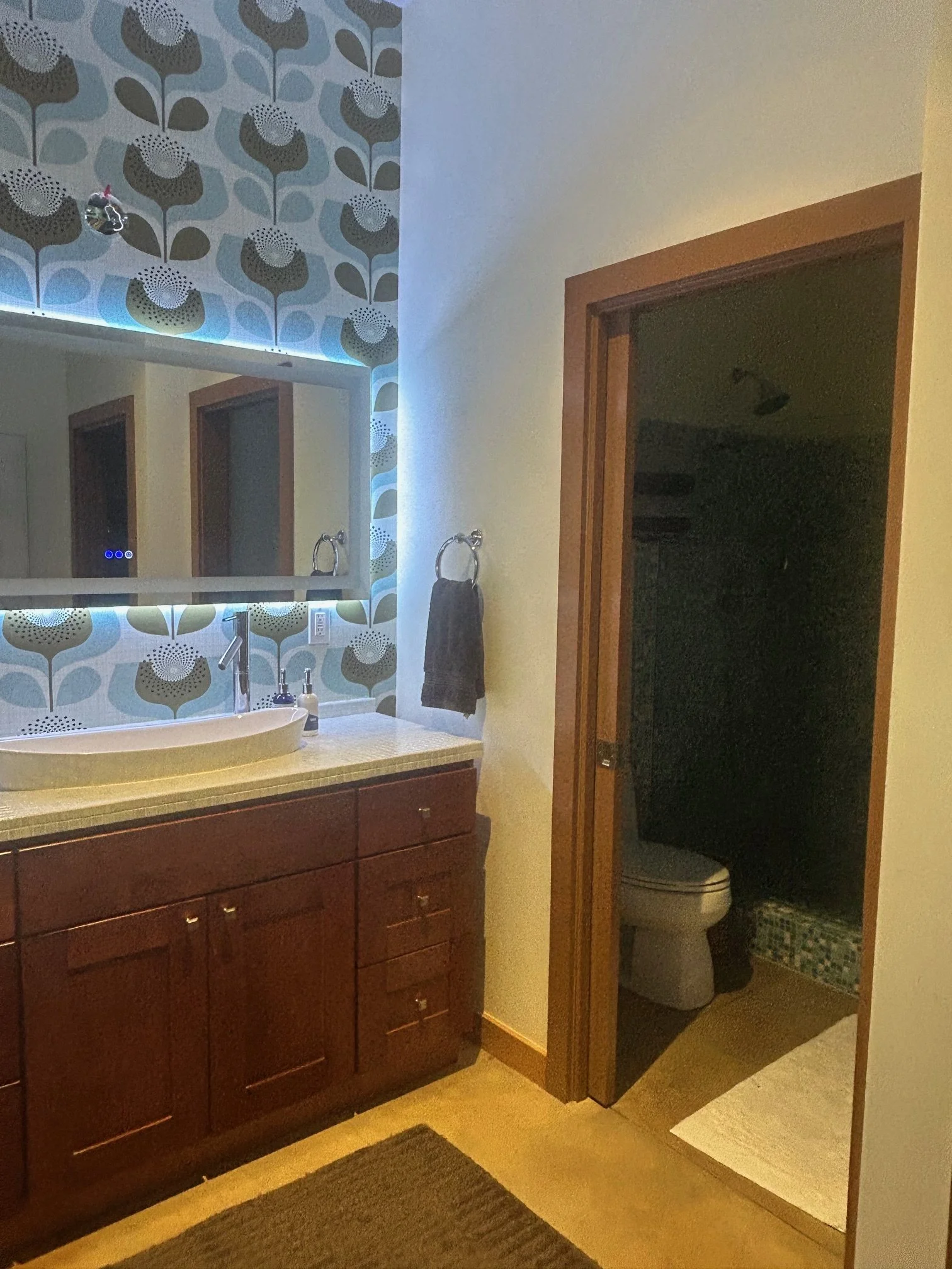 A bathroom with a decorated wall featuring a floral pattern, a white sink with a wooden cabinet below, a mirror, a towel ring with a black towel, and a doorway leading to a small room with a toilet and a trash can.
