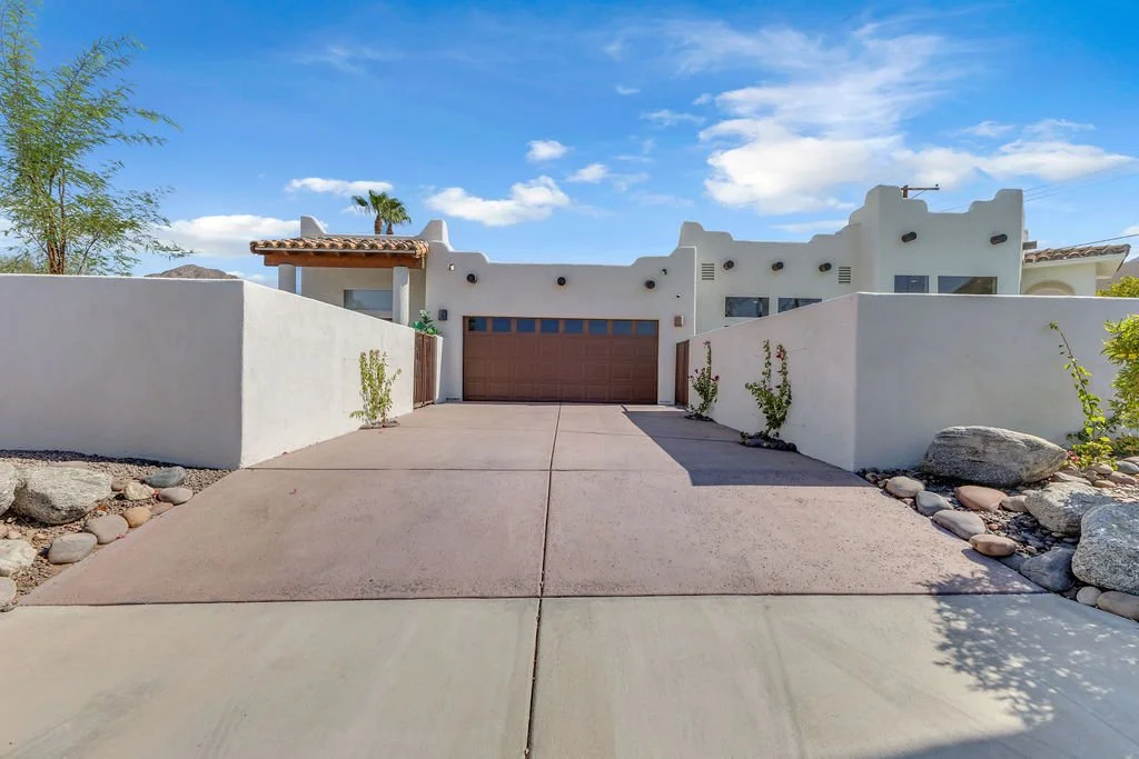 Front view of a modern white house with a brown garage door, desert landscaping, and a clear blue sky.