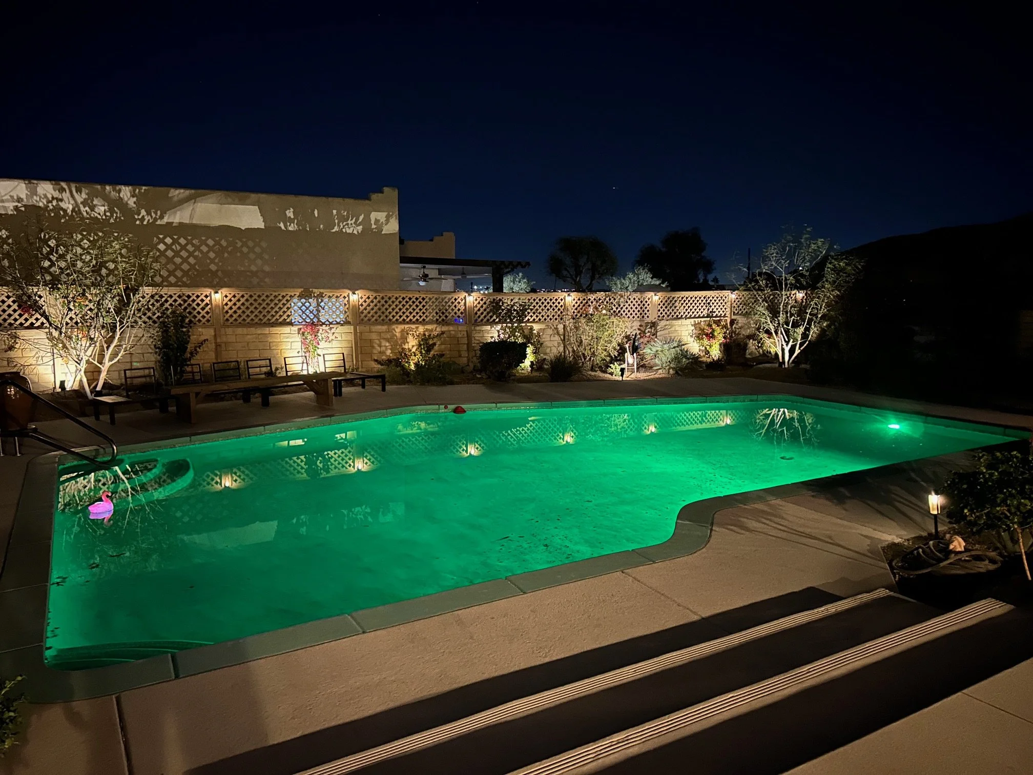 Nighttime backyard scene featuring a lit swimming pool with green lighting, surrounded by a concrete deck, wooden benches, trees, the fence with lights, and a clear view of the dark sky with a few visible stars.