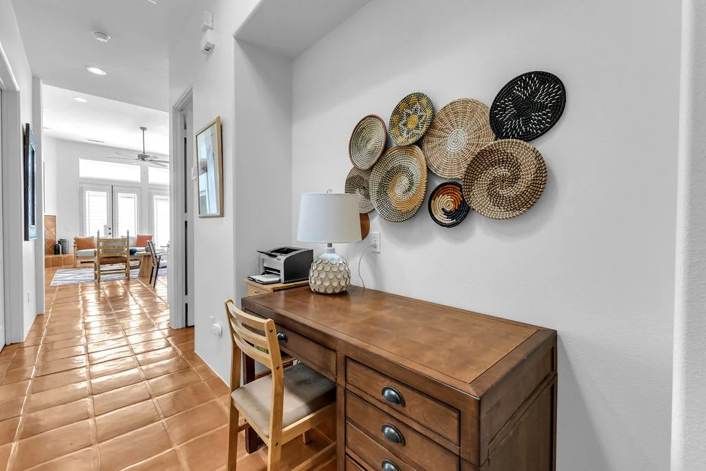 A hallway with terracotta tile floor, leading to a dining area with windows, chairs, and a table. The hallway features a wooden desk with a lamp, printer, and papers, and a wooden chair. Decorative woven baskets hang on the white wall above the desk.
