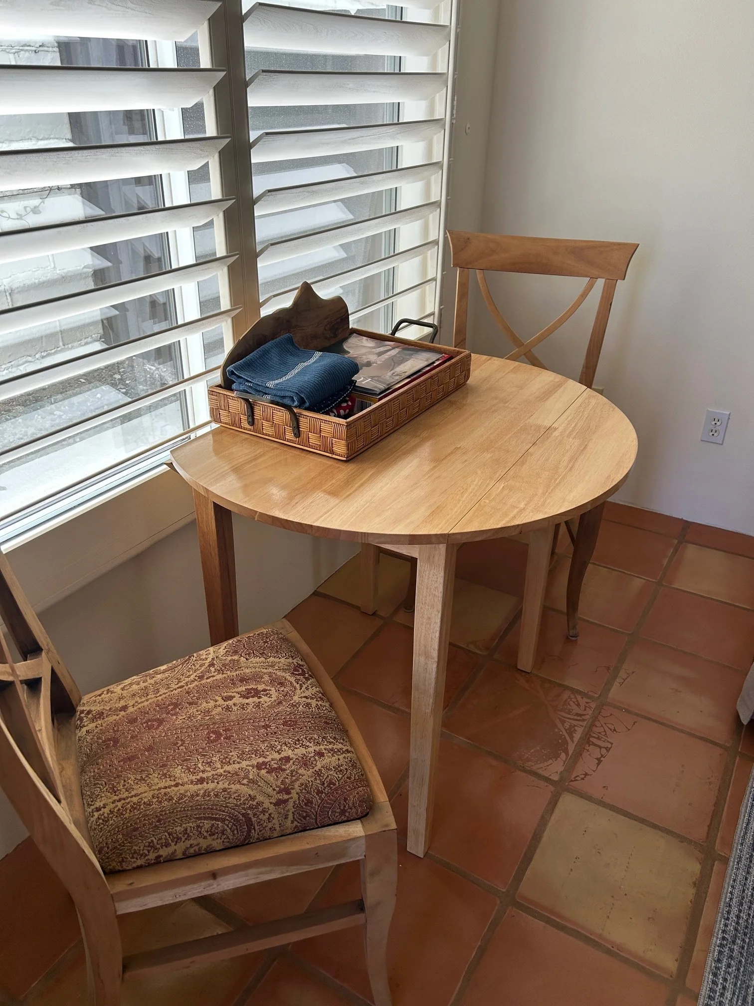 A dining room corner with a wooden table, one wooden chair with a decorative cushion, and another wooden chair without a cushion. There is a window with white plantation shutters and a windowsill. On the table, there is a wicker tray with a folded bl