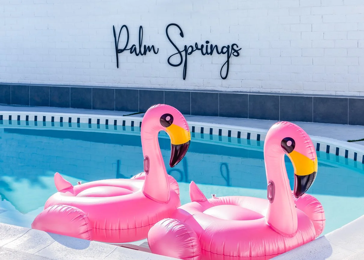 Two pink flamingo pool floats in a swimming pool with a white brick wall and a sign that says 'Palm Springs' in the background.