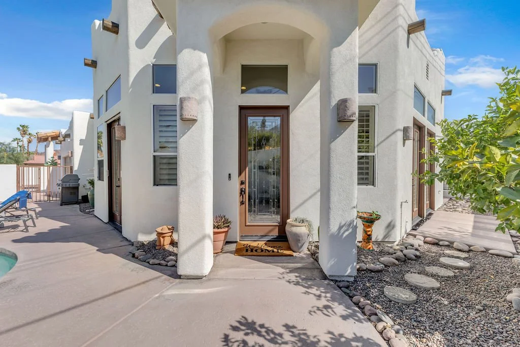 Front entry of a white stucco house with a glass door, potted plants, and a decorative mat that says 'Home'.