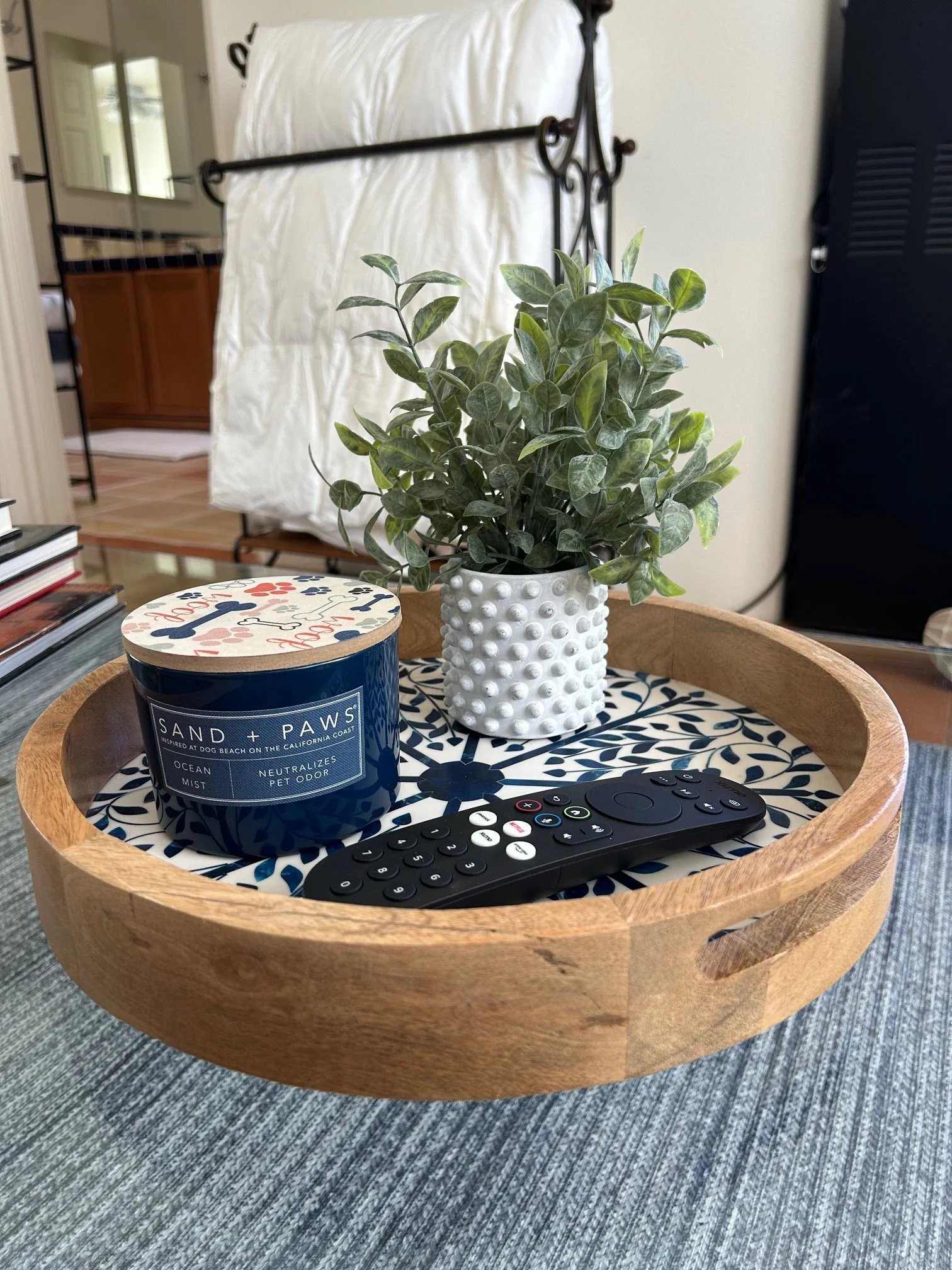 A round wooden tray with black and white patterned paper lining, holding a TV remote, a blue candle labeled "Sand + Paws" with dog-themed design on the lid, and a potted plant with green variegated leaves in a white textured pot. The tray is on a blu