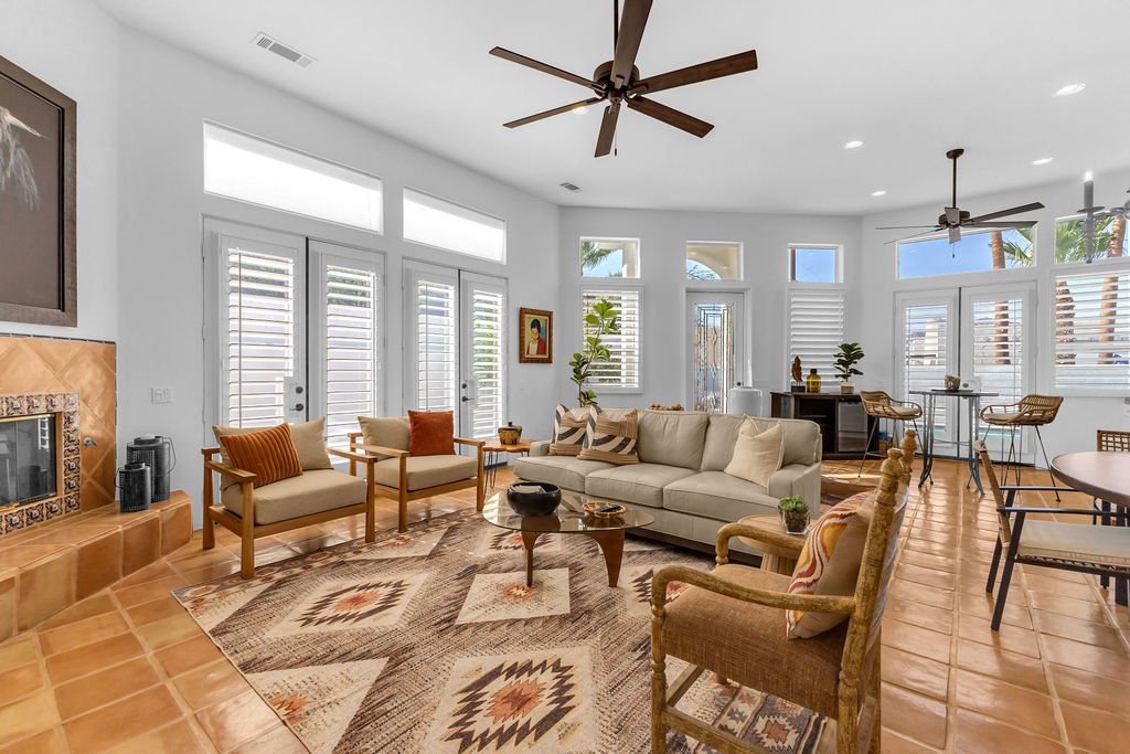 Bright living room with white walls, wooden furniture, a fireplace, and a large window with shutters.