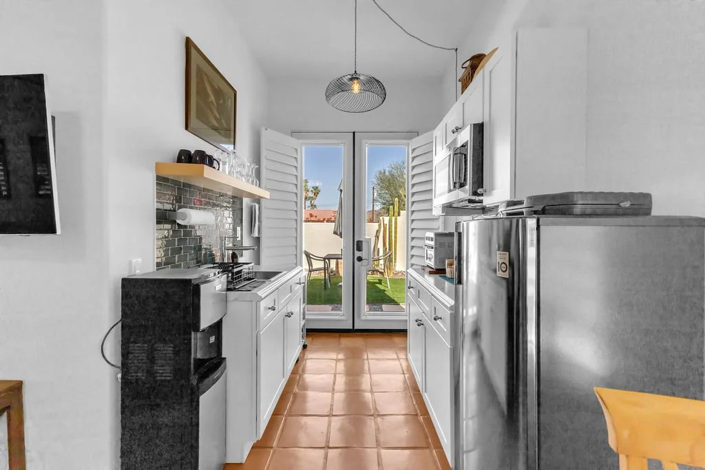 Small kitchen with white cabinets, stainless steel appliances, a black coffee machine, and a sliding glass door leading to an outdoor patio with chairs and a view of trees and neighboring houses.
