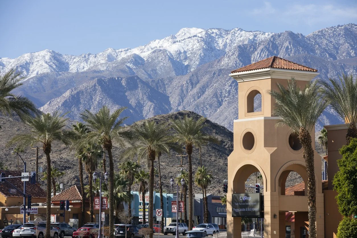A small town with palm trees, a clock tower, and snow-capped mountains in the background.