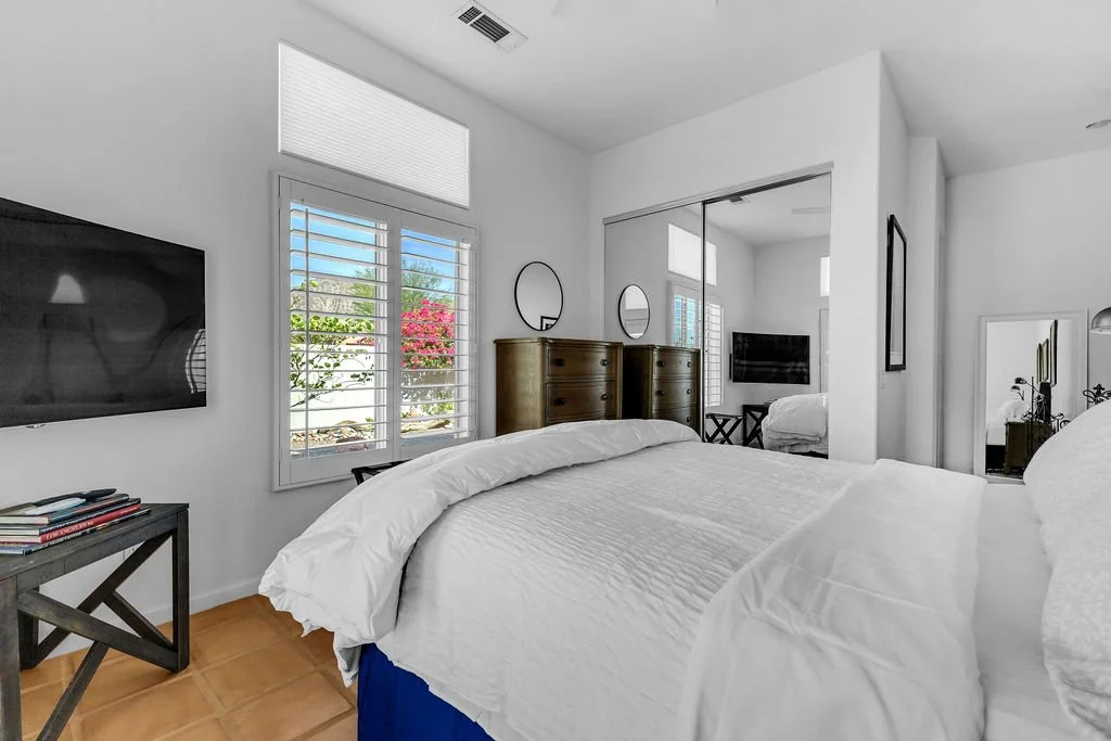 A bedroom with a white bed, a window with blinds, a flat-screen TV mounted on the wall, a small side table with books, wooden dressers with mirrors, and a sliding closet door with a mirror.