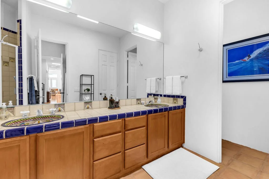 A bathroom with a wooden vanity, tiled countertop with colorful design sinks, a large mirror, and tropical decor elements.
