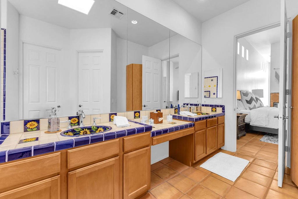 A bathroom with a double vanity with sunflower patterned blue and yellow tiles, wooden cabinets, a large mirror, and a view into a bedroom with a bed, nightstand, and window.