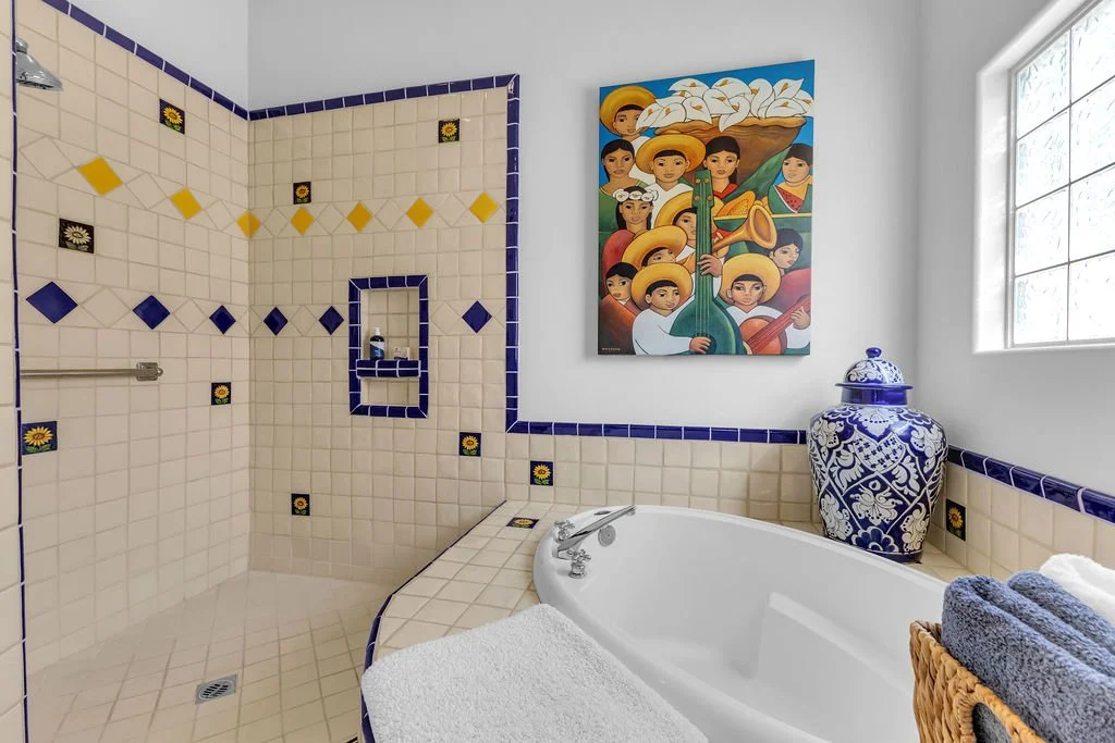 Bathroom with a tiled shower area, a white bathtub with a towel, a large blue and white decorative jar on the ledge, a colorful mural of Mexican musicians, and a window with frosted glass blocks.