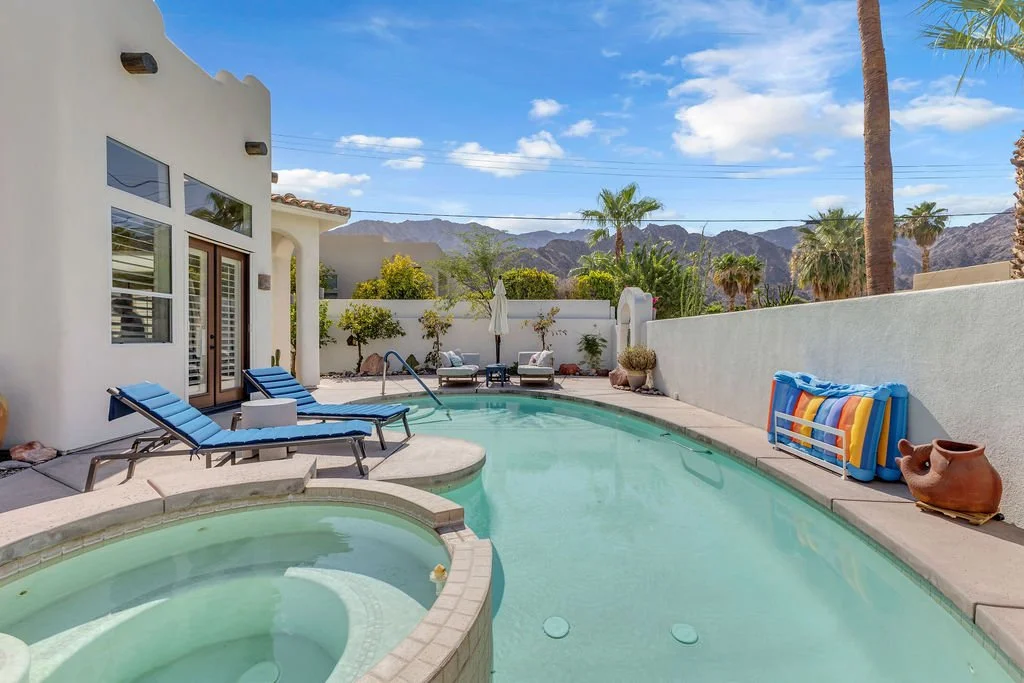 A backyard with a swimming pool, lounge chairs, potted plants, and mountain views under a blue sky.