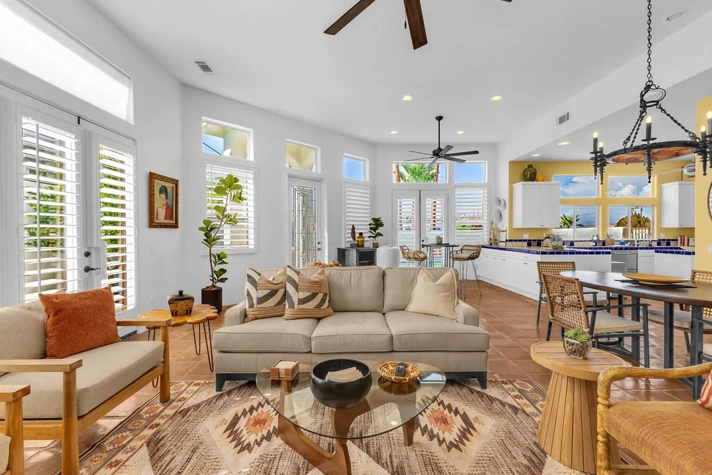 Open-concept living room and kitchen with white walls, multiple windows with shutters, beige sofa, wooden chairs, modern decor, and a tiled floor.