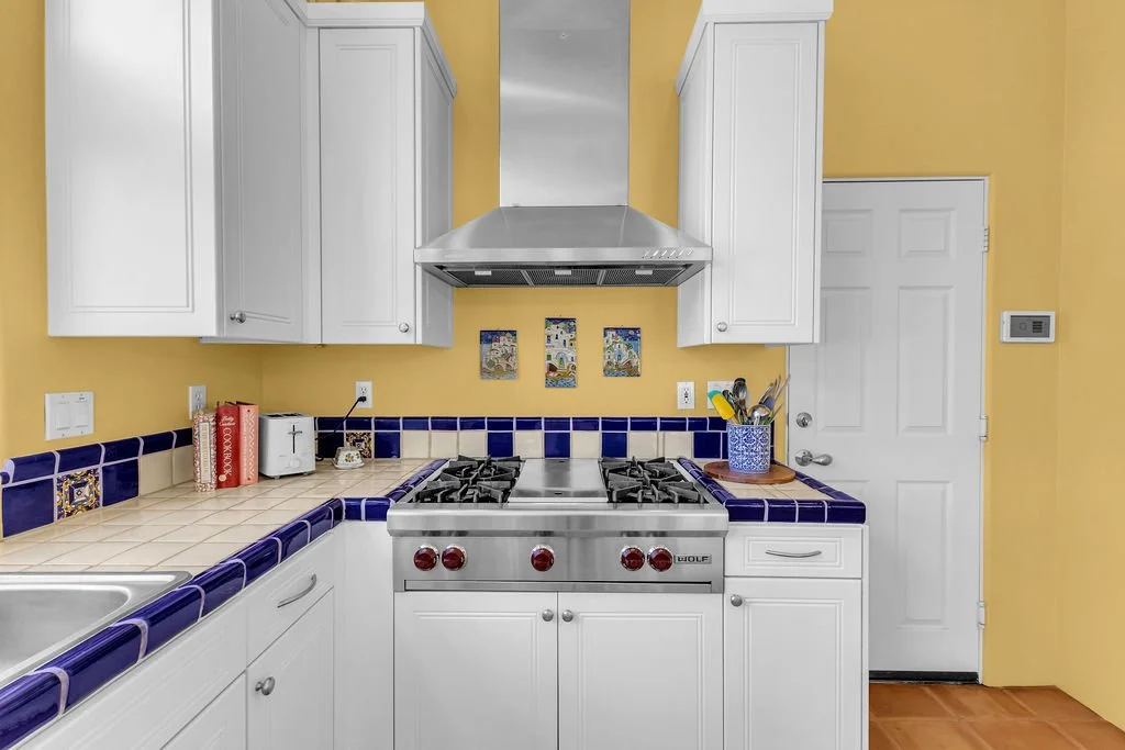 Kitchen with yellow walls, white cabinets, a range with a stainless steel hood, blue and white tiled countertops, and a white door.