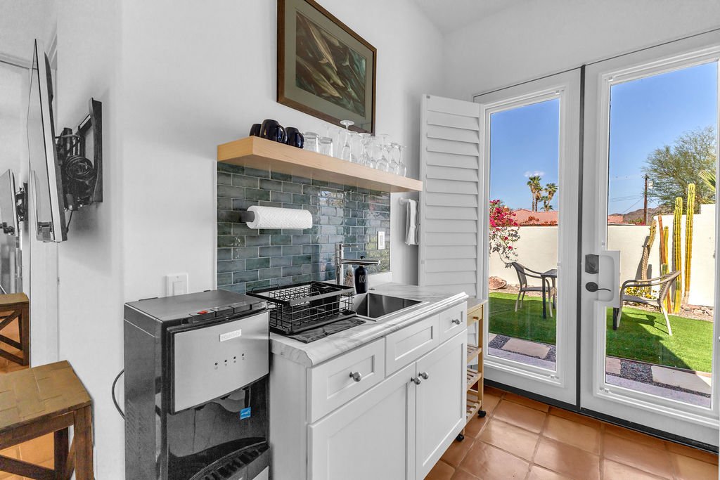 Small kitchen with white cabinets, a sink, and a countertop, leading to an outdoor patio with chairs and tall cactus plants, through large glass doors.