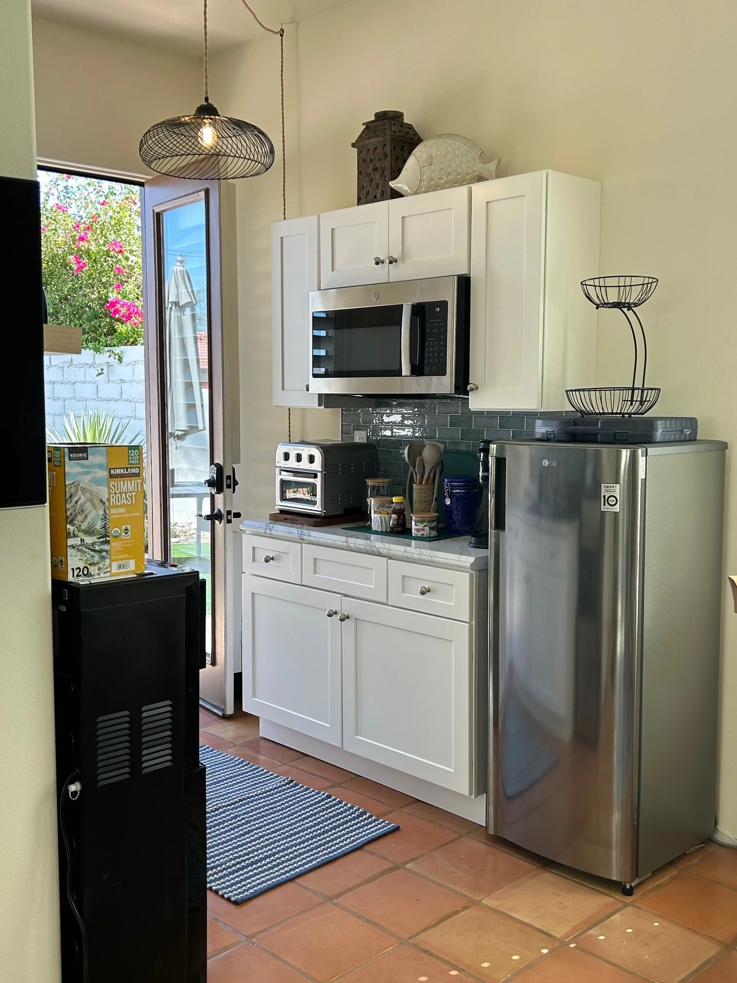 A kitchen corner with white cabinets, a microwave, a stainless steel refrigerator, and a small kitchen counter with various items. There is a doorway to the outside with a view of pink flowering plants and a white umbrella. Overhead, a black wire pen