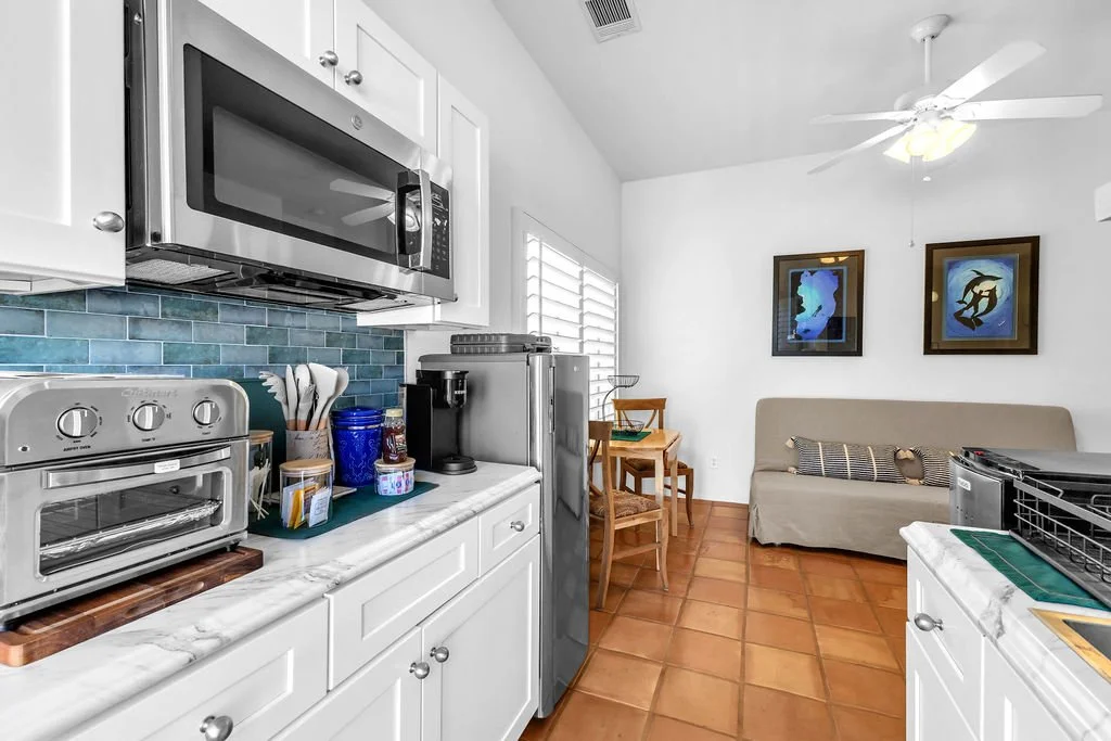 Kitchen and living area with white cabinets, a microwave, oven, and coffee machine on the counter, a window with blinds, two framed artworks on the wall, a small beige sofa with pillows, a ceiling fan, and a tiled floor.