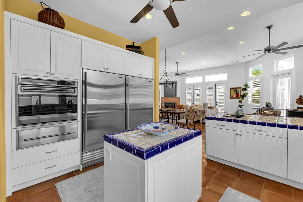 Open-concept kitchen with white cabinets, stainless steel appliances, and a small island with blue and white tiled top, overlooking a living room with large windows and seating area.