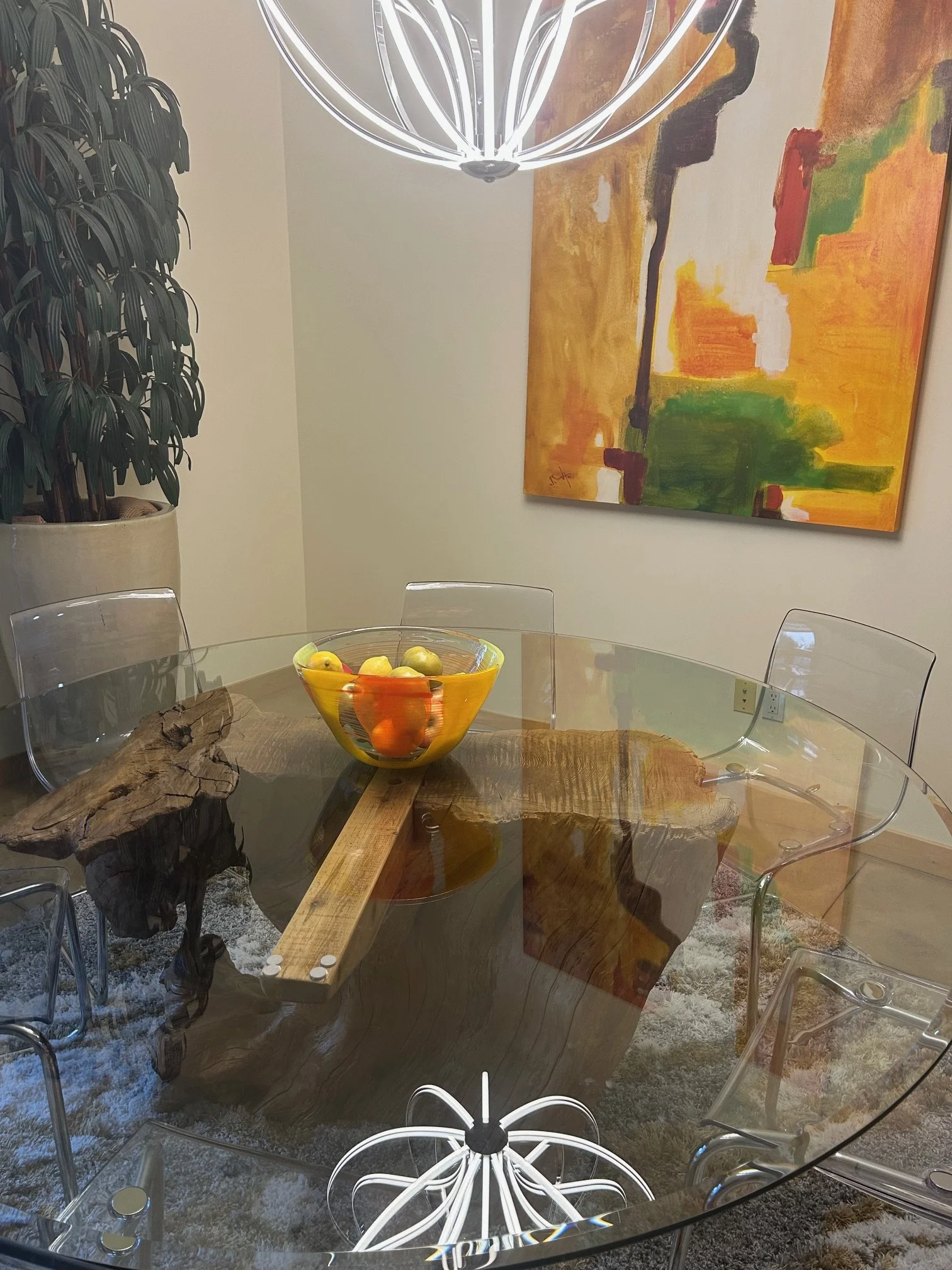 A modern dining room with a glass table featuring a wooden centerpiece, a bowl of mixed fruits, a large abstract painting with warm colors on the wall, a green plant in a beige pot, and a contemporary metal light fixture above the table.