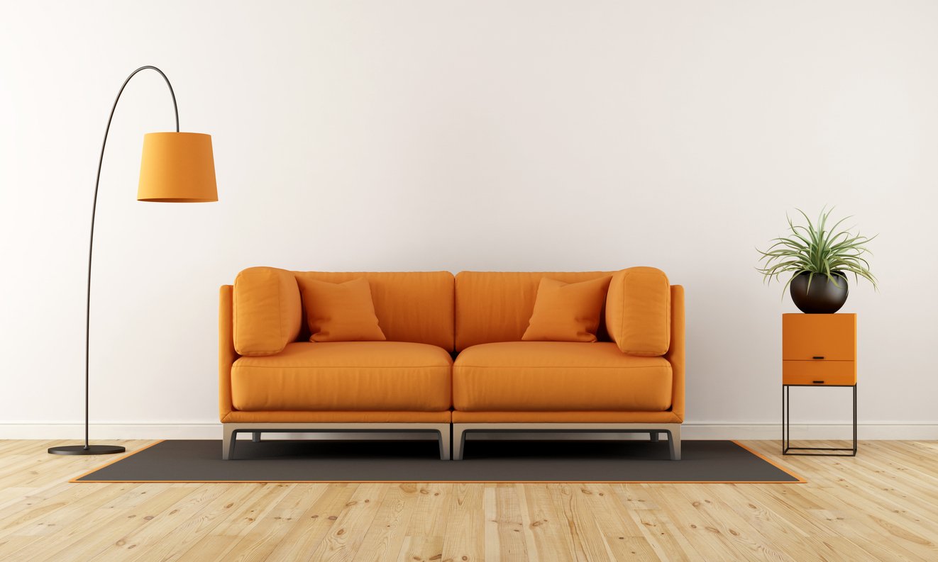 An orange sofa with three cushions, a black and orange side table with a plant, an orange floor lamp, on a gray rug, in a minimalist room with a white wall and wood flooring.