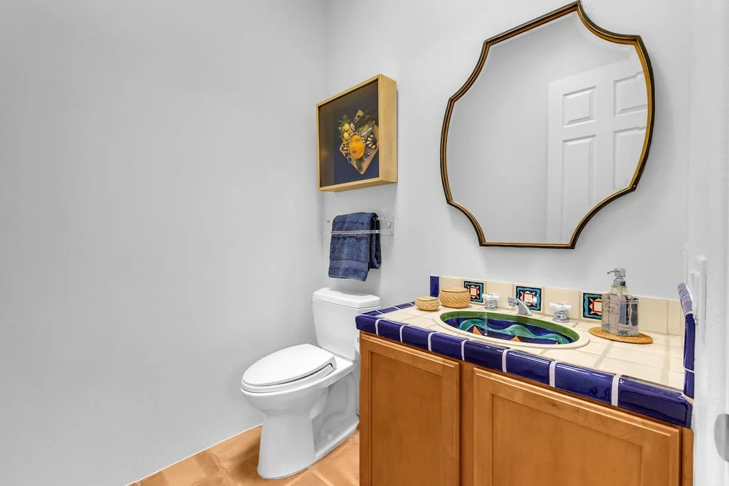 Bathroom with a toilet, wooden cabinet with a dark blue tiled countertop, decorative sink, mirror, towel rack with a dark blue towel, and wall art with a floral arrangement.