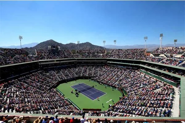 A large outdoor tennis stadium filled with spectators during a match on a sunny day with mountainous terrain in the background.