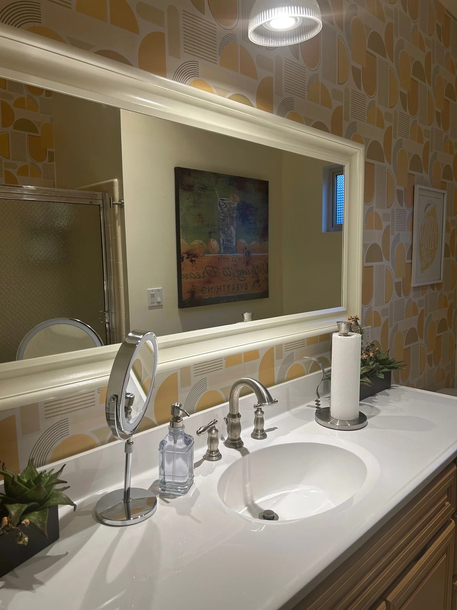 A bathroom vanity with a white countertop, oval sink, and silver faucet. A roll of paper towels and small plants are on the counter. A large mirror above reflects a framed art piece on a wall with geometric patterned wallpaper. The reflection in the 