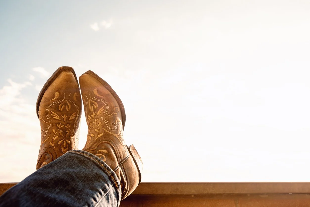 Person wearing cowboy boots with intricate designs, sitting and relaxing with legs crossed on a wooden surface, looking up at the sky.