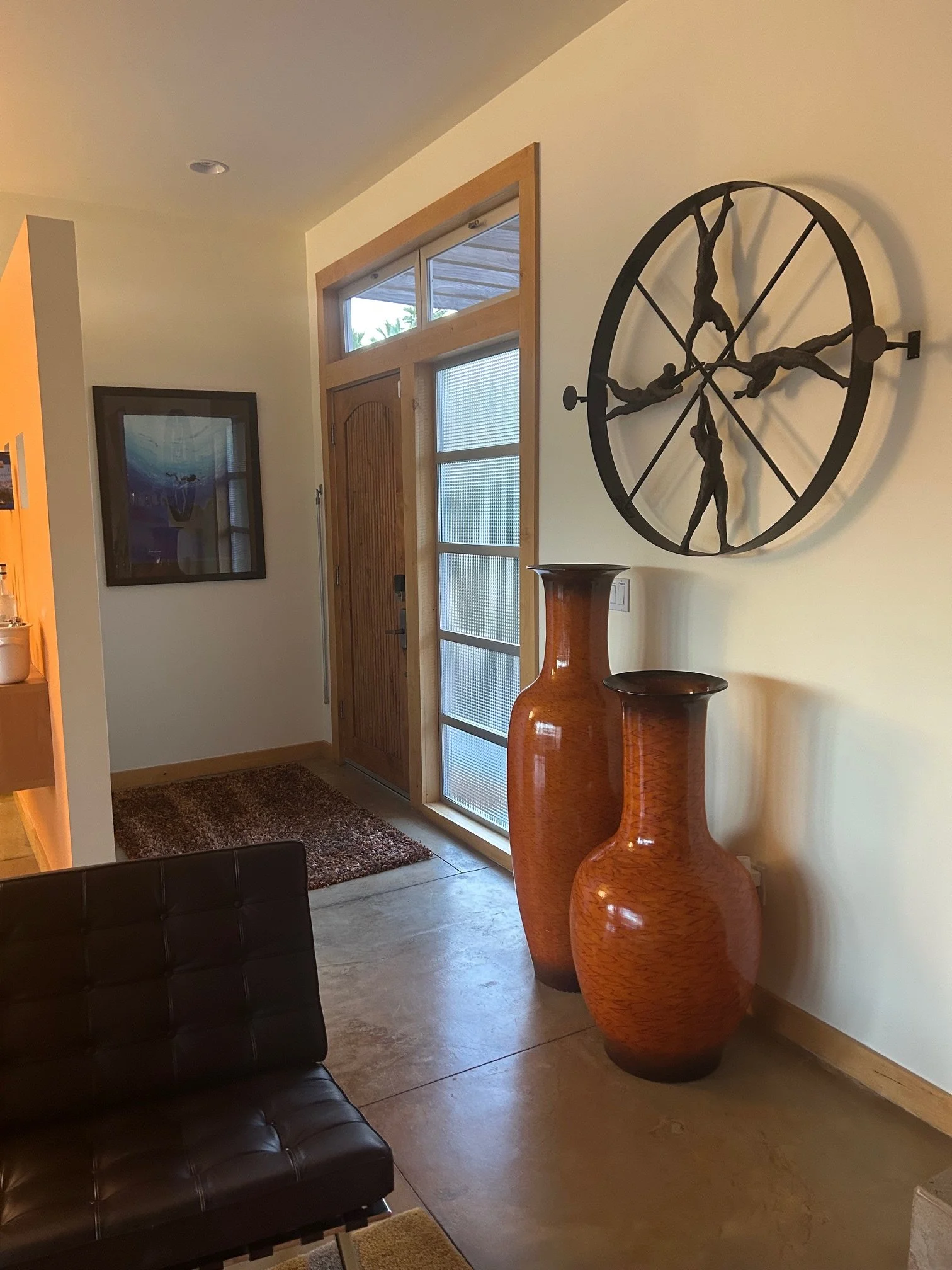 Living room entrance with wooden door, two large orange vases, a wall clock with human figures in dynamic poses, a framed picture on the wall, a brown leather couch, and a small wooden table.