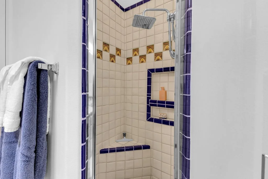 A small tile shower with purple border tiles and a built-in ceramic shelf with lotion bottle and soap bar, with towels hanging outside.