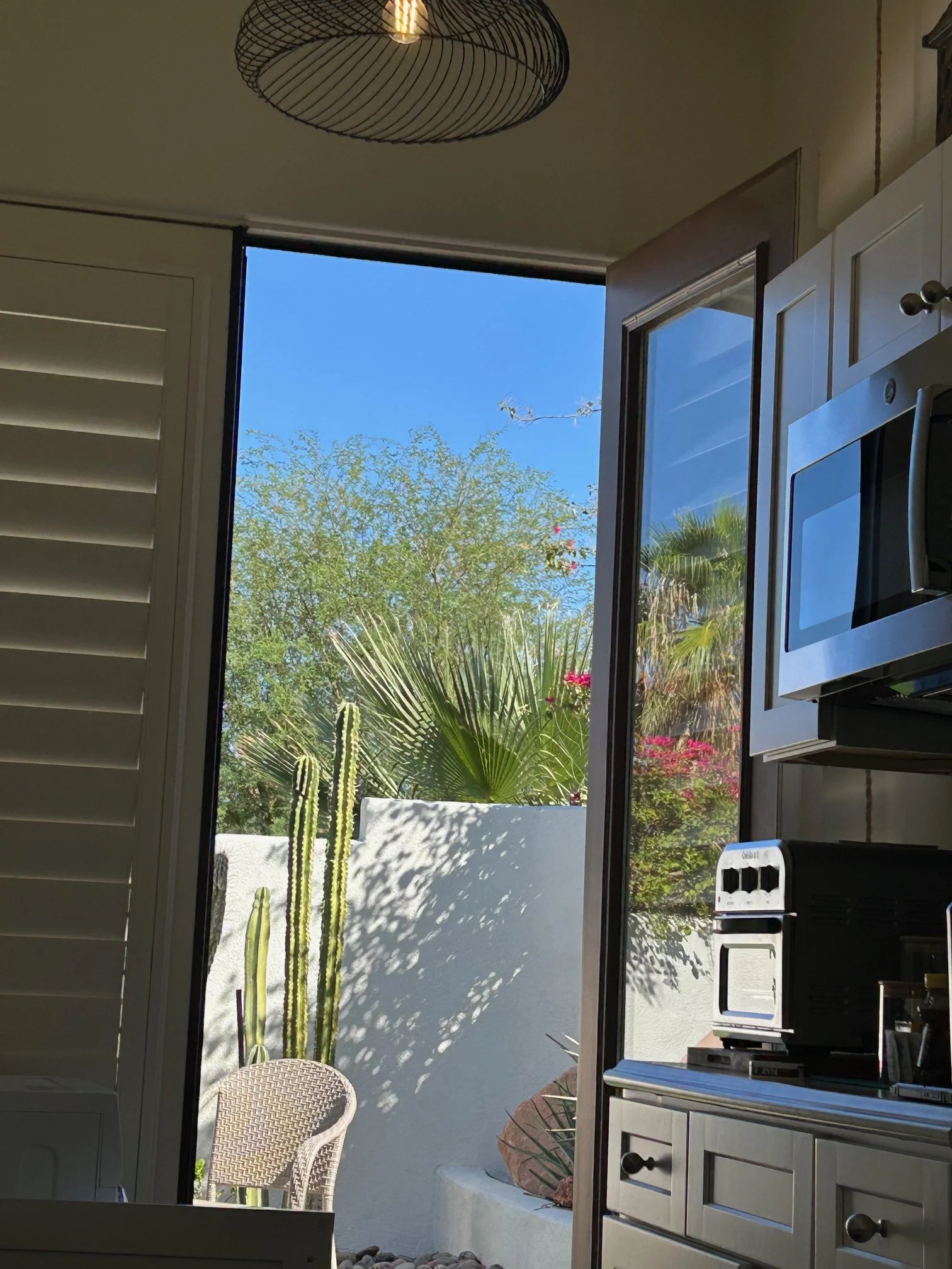 View from inside a kitchen looking out through a large open window to a sunny outdoor patio with tall cacti, a palm tree, and other desert plants, with clear blue sky.