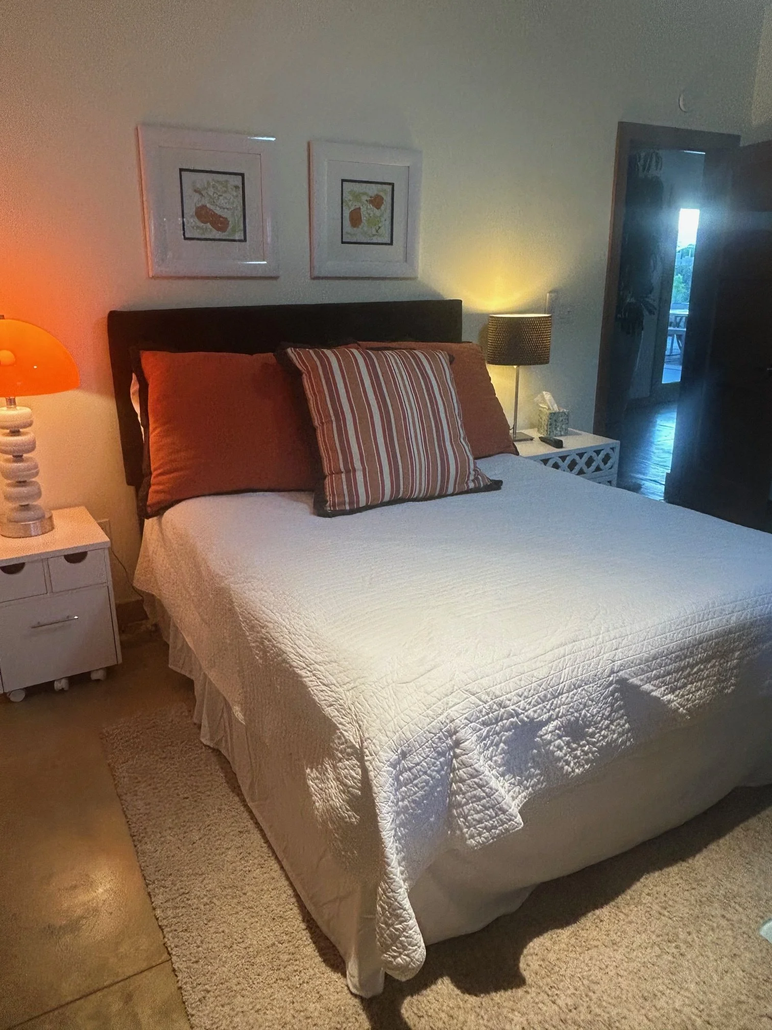 A neatly made bed with a white quilt, large striped pillow, and dark wooden headboard. There are two framed botanical art prints above the bed. To the left, there is a white nightstand with a decorative lamp with an orange shade. To the right, there 