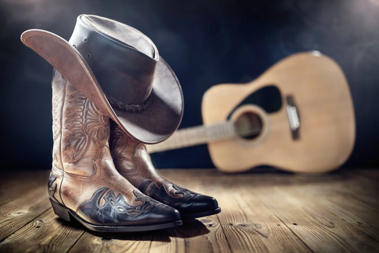 A pair of cowboy boots with intricate stitching, a leather cowboy hat, and a wooden acoustic guitar on a wooden surface with a dark background.