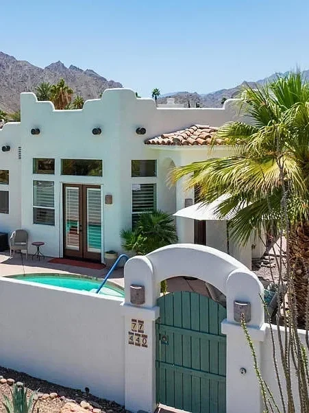 A white southwestern-style house with a small swimming pool, palm trees, and mountains in the background.