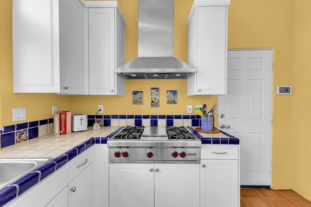 A kitchen with white cabinets, a yellow wall, and blue-tiled countertops. There is a stainless steel stove with a range hood, a white door, and a small white refrigerator on the counter. The counter has a few books, a toaster, a cup, and a blue conta