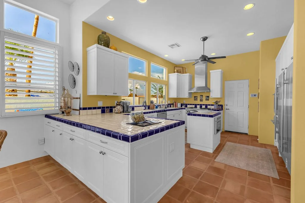 Bright yellow kitchen with white cabinets and blue tiled countertops. Window with white blinds, ceiling fan, and terracotta tiled floor. Outside palm trees visible through windows.