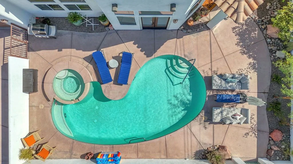 An aerial view of a backyard with a swimming pool, hot tub, two blue lounge chairs, a small white table, and other outdoor furniture, surrounded by plants and patio flooring.