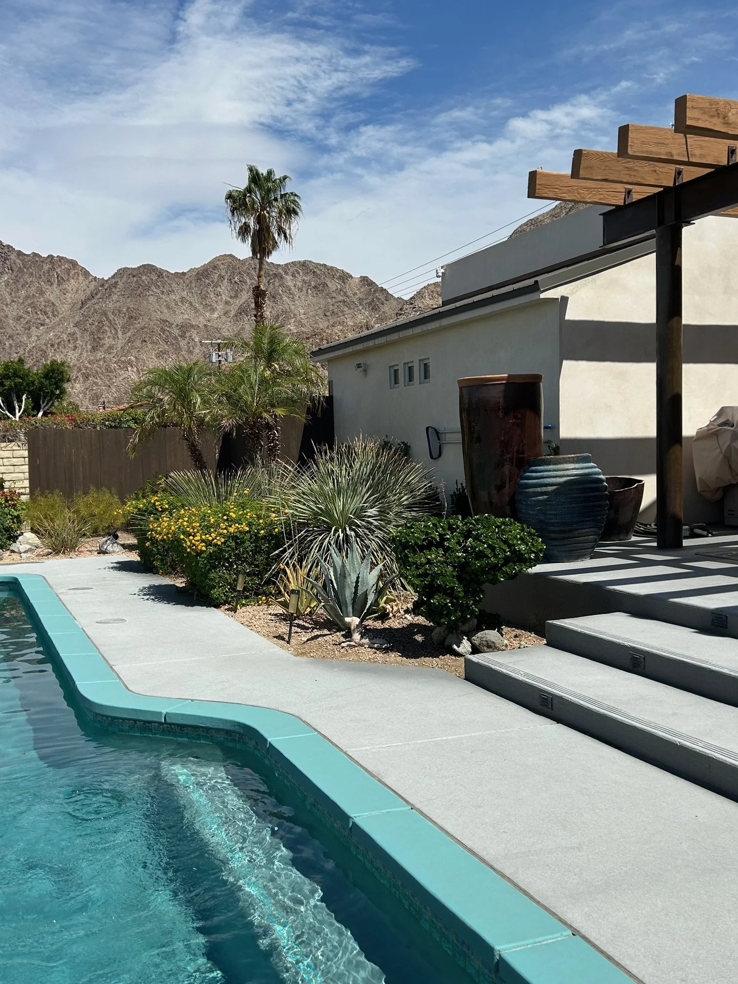 A swimming pool surrounded by a patio with potted plants, stairs, and a white building. In the background, there are dry, rugged mountains and a partly cloudy sky.