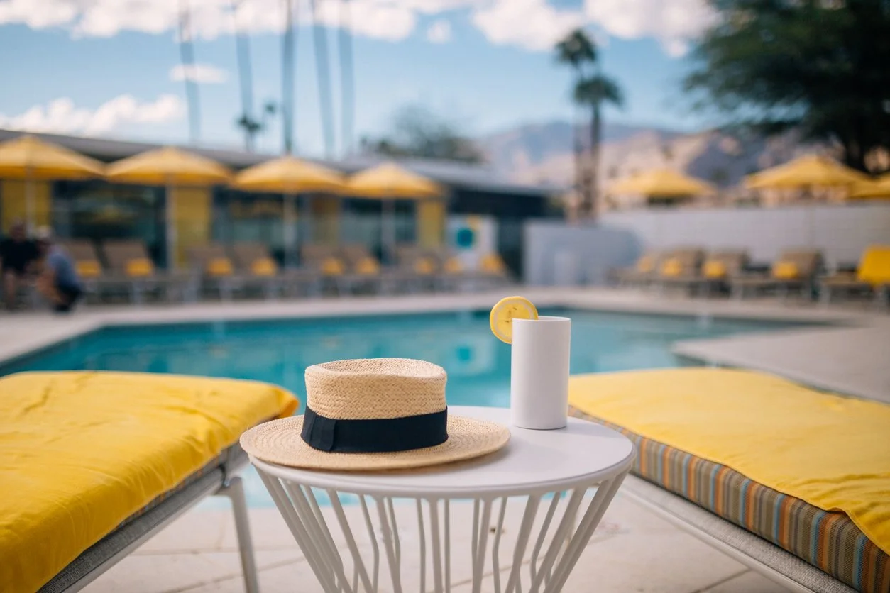 Poolside scene with yellow lounge chairs, a straw hat and a drink with lemon slice on a small white table, overlooking a swimming pool with yellow umbrellas and palm trees in the background.