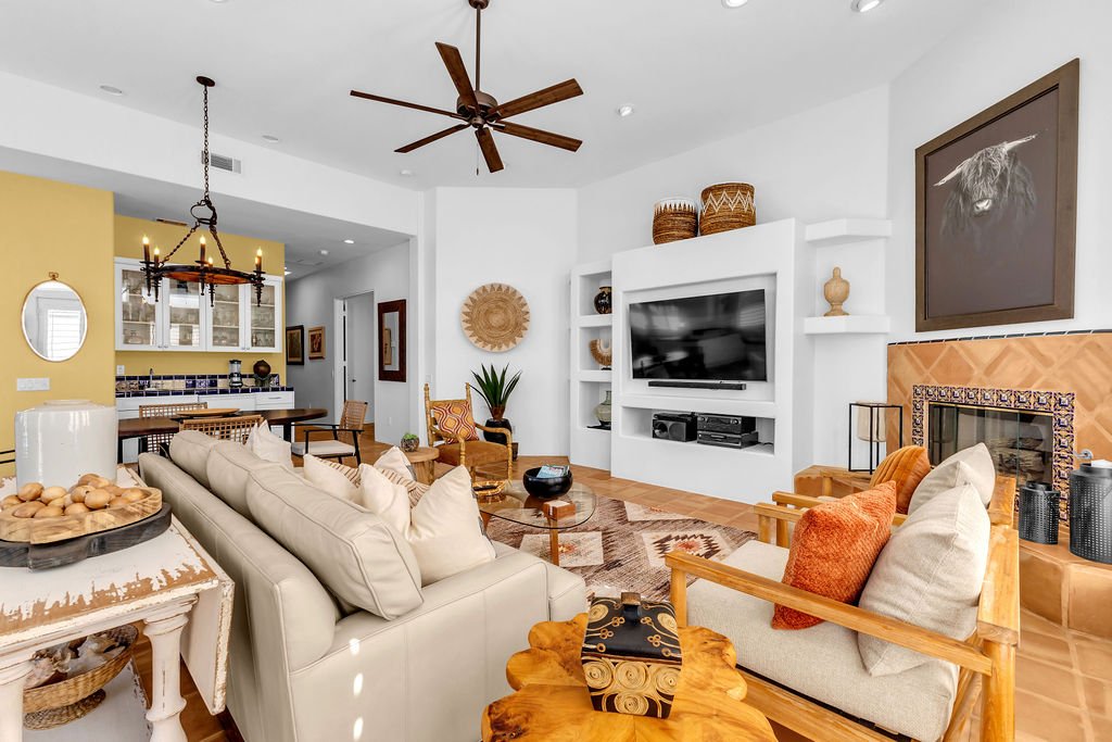 Living room with a beige sofa, wooden armchair, and a fireplace, with a flat-screen TV on white built-in shelves, artwork on the wall, and a ceiling fan.