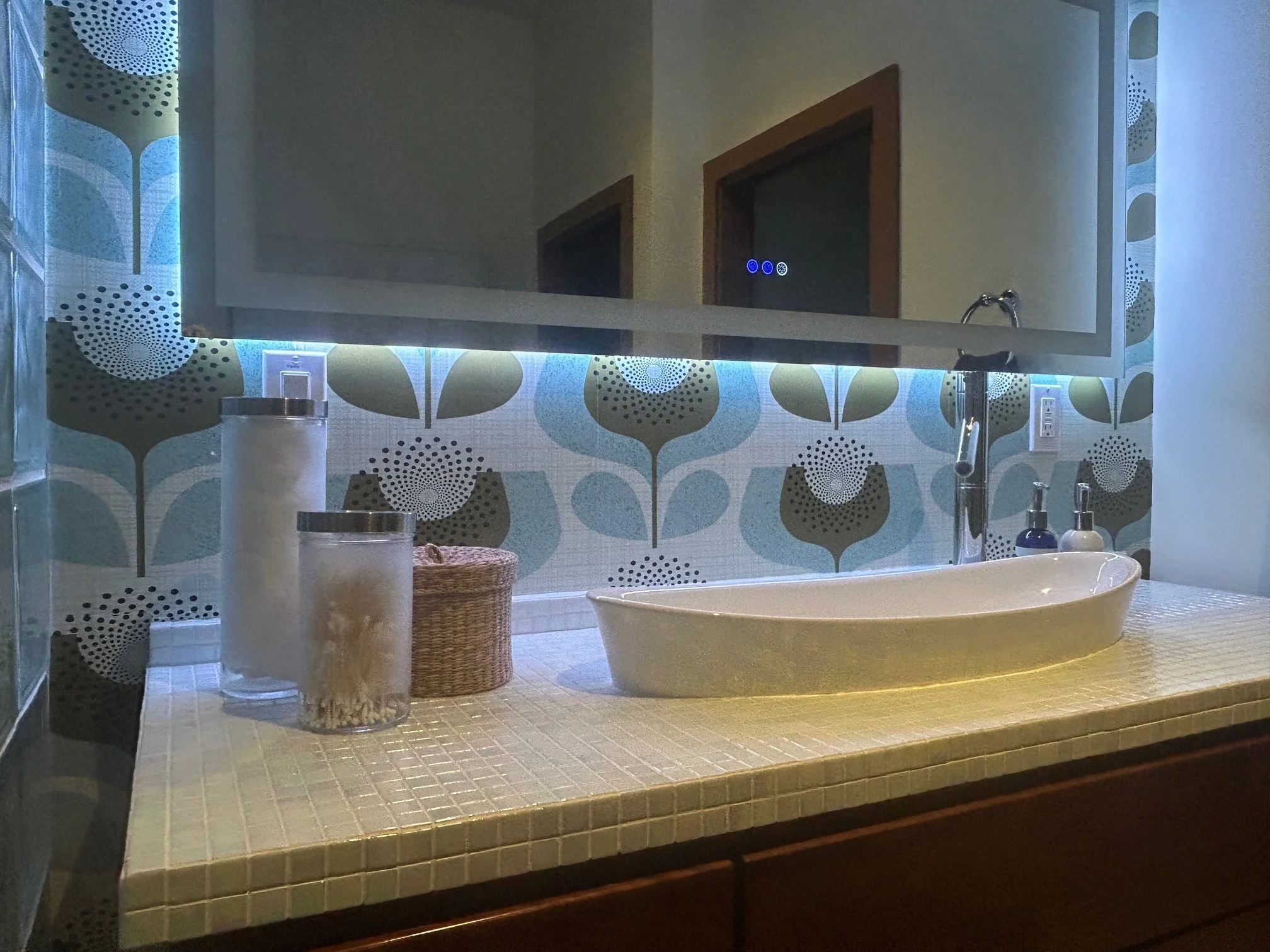 A bathroom with a tiled countertop, a mirror with blue backlighting, and a sink with a faucet. There are bottles, jars, and a basket on the countertop. A wallpaper with a leaf pattern decorates the wall behind the mirror.