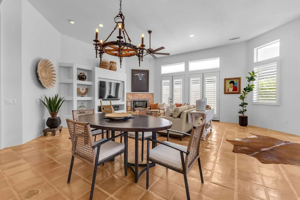 Bright living room with white walls, a dark wood dining table with four chairs, a beige sofa, a fireplace, and large windows with shutters. Decor includes plants, artwork, and a chandelier.