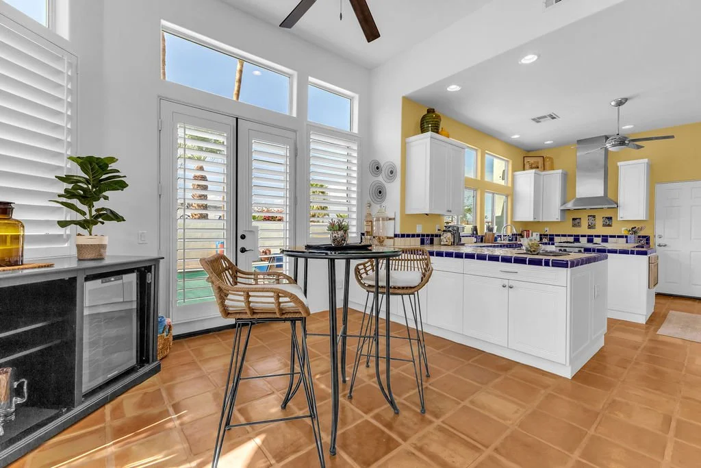 Bright kitchen with white cabinets, yellow wall, purple tiled countertop, and terracotta tile floor. Includes breakfast bar with two wicker chairs, a black sideboard with potted plant, and large glass-door with valances and window shutters.